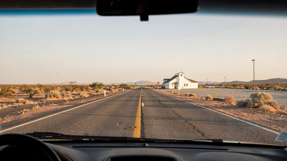 A long two-lane desert road at late afternoon, seen from the passenger seat of a car, with a small white church and a gravel parking lot visible in the distance under a wide sky.